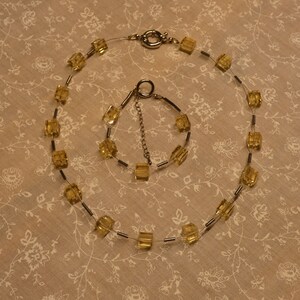 Könnte beinhalten: Ein Halsketten- und Armband-Set mit quadratischen, bernsteinfarbenen Perlen. Die Halskette und das Armband sind auf einem dünnen, transparenten Draht mit silberfarbenen Metallakzenten aufgefädelt. Die Halskette hat einen silbernen Verschluss. Das Armband hat eine Kette zur Größenanpassung.