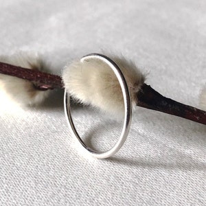 May include: A simple silver ring with a smooth, rounded surface. The ring is shown resting on a white, fuzzy surface.
