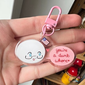 May include: A clear acrylic keychain with a pink clasp. The keychain features a white cartoon character with blue eyes and a tongue sticking out, and a pink speech bubble that says "you're a dumb whore" in red text. A small American flag is also attached.