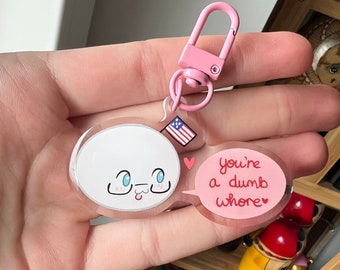 Puede incluir: Un llavero acrílico transparente con un cierre rosa. El llavero presenta un personaje de dibujos animados blanco con ojos azules y una lengua afuera, y un bocadillo rosa que dice "you're a dumb whore" en texto rojo. También hay una pequeña bandera estadounidense adjunta.