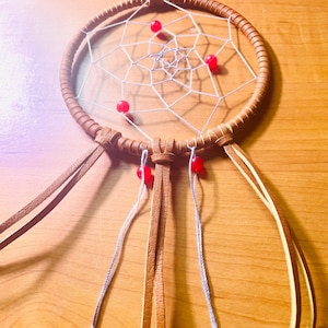 May include: A handcrafted dreamcatcher with a brown leather ring and woven web. Red beads are evenly spaced around the web and on the hanging leather strands. The dreamcatcher is designed to be hung.