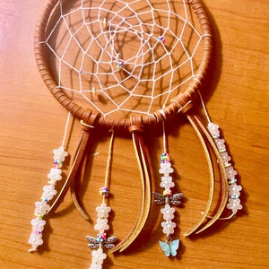 May include: A brown leather-wrapped dreamcatcher with a white web design. The dreamcatcher has hanging strands with white flower beads, dragonfly charms, and light blue butterfly charms. The dreamcatcher is on a wooden surface.