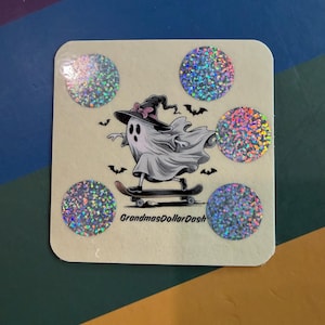 May include: A square sticker featuring a ghost wearing a witch hat and riding a skateboard, surrounded by small bats. The design includes four holographic circular accents and the text GrandmasDollarDash printed in black at the bottom center.