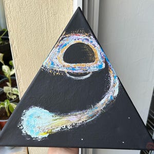 May include: A triangular canvas painting featuring a black background with a central dark circular void surrounded by a swirling ring of textured paint in shades of blue, gold, white, and purple, extending into a comet-like tail at the bottom.