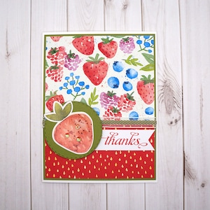 May include: A handmade greeting card with a watercolor design of strawberries, blueberries, and raspberries. The card has a green circle with a strawberry in the center and the word "thanks" written in red on a white banner.