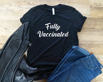 May include: Black t-shirt with white text that reads "Fully Vaccinated". The shirt is laid flat on a wooden surface with a leather jacket and blue jeans beside it.
