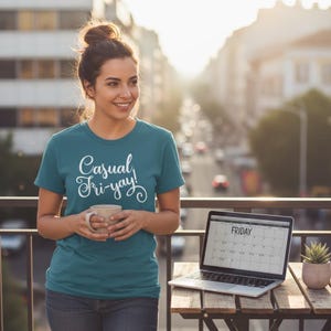 Puede incluir: Camiseta de color verde azulado con el texto "Casual Fri-yay!" en escritura blanca. Una mujer sostiene una taza y sonríe. Un portátil que muestra un calendario con "Viernes" resaltado está sobre una mesa de madera. También hay una pequeña planta en maceta sobre la mesa.
