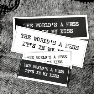 以下が含まれることがあります： 「The world's a mess, it's in my kiss」というテキストが書かれた黒と白のステッカーが4枚。ステッカーはデニムの背景に置かれています。