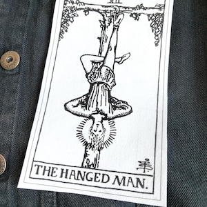 May include: A white rectangular patch featuring a black and white illustration of The Hanged Man tarot card. The image depicts a figure hanging upside down, with the text "THE HANGED MAN" below. The patch is set against a dark denim background.