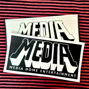 以下が含まれることがあります： 太字の白い3Dスタイルの文字で「MEDIA」という単語が書かれた2つの長方形のステッカー。1つのステッカーは黒い背景で、もう1つは白い背景です。黒いステッカーの「MEDIA」という単語の下には「MEDIA HOME ENTERTAINMENT」というテキストがあります。