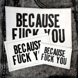 May include: Three rectangular white fabric patches of varying sizes, each featuring the black printed text "BECAUSE FUCK YOU" in a bold, sans-serif font. The patches are layered on top of each other against a dark grey acid-wash denim fabric background.