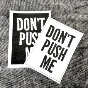 Könnte beinhalten: Zwei schwarz-weiße Schilder mit dem Text "Don't Push Me" darauf gedruckt. Die Schilder befinden sich auf einem grauen Jeans-Hintergrund.