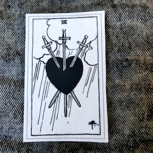 May include: Black and white tarot card featuring three swords piercing a black heart. The card is labeled "III" at the top and depicts a stormy sky with rain. A small symbol is at the bottom.