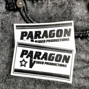 Puede incluir: Dos etiquetas rectangulares blancas con el texto "PARAGON VIDEO PRODUCTIONS" en negro. Una etiqueta tiene un gráfico de estrella. Las etiquetas están sobre un fondo de mezclilla gris.