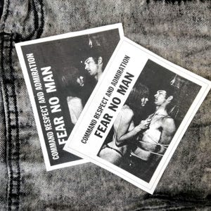 May include: Two black and white stickers with the text "COMMAND RESPECT AND ADMIRATION FEAR NO MAN" and an illustration of a couple. The stickers are on a grey denim background.