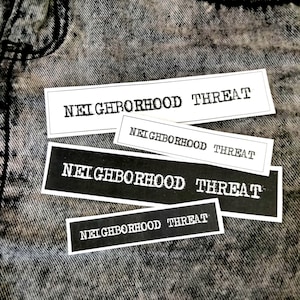 Peut inclure: Quatre autocollants rectangulaires avec le texte "NEIGHBORHOOD THREAT" en gras, en police sans-serif. Deux autocollants sont blancs avec du texte noir, et deux sont noirs avec du texte blanc. Les autocollants sont disposés sur une surface en denim gris texturé.
