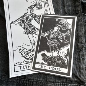 May include: Two black and white tarot card patches featuring "The Fool" card. The cards depict a figure with a staff, sun, and mountains. The text "THE FOOL" is printed on the bottom of one card, and "THE" on the other.