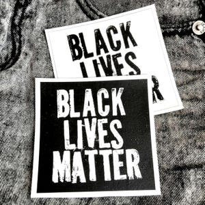 May include: Two square stickers with the text "BLACK LIVES MATTER" in bold, white letters. One sticker has a black background, and the other has a white background. The stickers are on a grey denim surface.