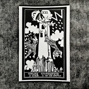 May include: Black and white tarot card depicting The Tower, card number XVI. The card shows a tall tower with a lightning bolt striking it. Two figures fall from the tower. The text "THE TOWER." is printed at the bottom of the card.