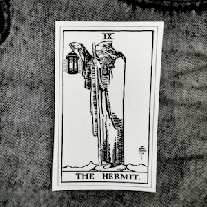 Op de afbeelding: Zwart-witte illustratie van de tarotkaart De Kluizenaar. De kaart toont een figuur in een gekapte mantel die een lantaarn en een staf vasthoudt. Het kaartnummer IX staat rechtsboven. De tekst "DE KLUIZENAAR" staat onderaan de kaart.