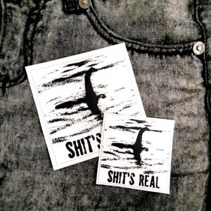 Könnte beinhalten: Zwei weiße quadratische Aufkleber mit einer Schwarz-Weiß-Illustration eines Wesens im Wasser. Der Text "SHIT'S" befindet sich auf dem größeren Aufkleber und "SHIT'S REAL" auf dem kleineren. Die Aufkleber befinden sich auf einem grauen Jeans-Hintergrund.
