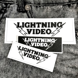 May include: Three stickers with the text "LIGHTNING VIDEO" in a bold, stylized font, accompanied by lightning bolt graphics. The stickers are white with black text and a black sticker with white text. The stickers are on a gray denim background.