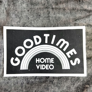 May include: A black rectangular sticker with the words "GOOD TIMES" in white, arched above a rainbow design, and "HOME VIDEO" below. The sticker is on a gray denim background.