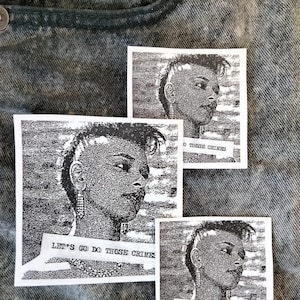 Op de afbeelding: Drie zwart-wit stickers met een portret van een persoon met een hanenkam en oorbellen. De tekst "LET'S GO DO THOSE CRIMES" staat op een banner onderaan elke sticker. De stickers liggen op een denim achtergrond.
