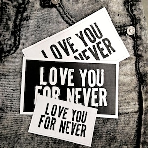 Könnte beinhalten: Drei rechteckige weiße und schwarze Karten mit dem Text "LOVE YOU FOR NEVER" in einer fetten, serifenlosen Schrift. Die Karten sind auf einer strukturierten, grauen Denim-Oberfläche angeordnet. Die Karten sind in verschiedenen Größen.