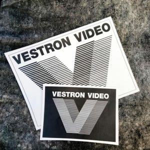May include: Two white and black Vestron Video stickers. The larger sticker has a white background with the text "VESTRON VIDEO" and a stylized "V" design. The smaller sticker has a black background with the same text and design.