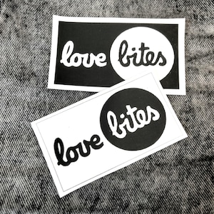 May include: Two white rectangular stickers with the words "love bites" in a handwritten font. The word "bites" is in a black circle. The stickers are on a gray textured surface.
