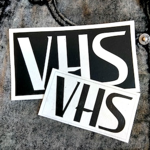 Pode incluir: Dois remendos retangulares brancos com a palavra "VHS" impressa em letras pretas. Os remendos estão sobre um fundo de sarja de algodão cinza escuro.