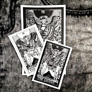 Könnte beinhalten: Schwarze und weiße Tarotkarten mit der Karte 'Die Liebenden' und anderen Designs. Die Karten zeigen detaillierte Illustrationen von Figuren und Symbolen. Sie liegen auf einer strukturierten Oberfläche.