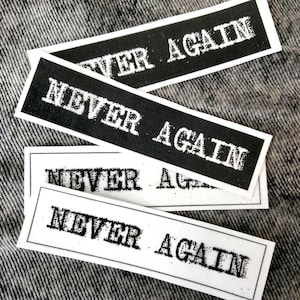 Puede incluir: Pegatinas rectangulares en blanco y negro con las palabras "NEVER AGAIN" impresas en una fuente audaz y desgastada. Las pegatinas están dispuestas sobre una superficie gris texturizada.