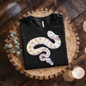 May include: A black short-sleeved t-shirt featuring a graphic print of a snake with a pale yellow and light purple patterned scale design. The snake is coiled in a curved shape across the center of the chest area of the garment.