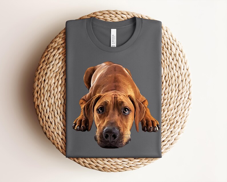 May include: A dark gray crew neck t-shirt featuring a detailed, realistic illustration of a brown Rhodesian Ridgeback dog lying down with its head facing forward and large, expressive eyes looking directly at the viewer.