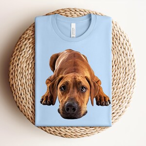 May include: A light blue t-shirt featuring a centered, detailed illustration of a brown dog in a lying position. The dog has large, expressive eyes, floppy ears, and a black nose. The shirt has a crew neckline and a white brand label at the neck.
