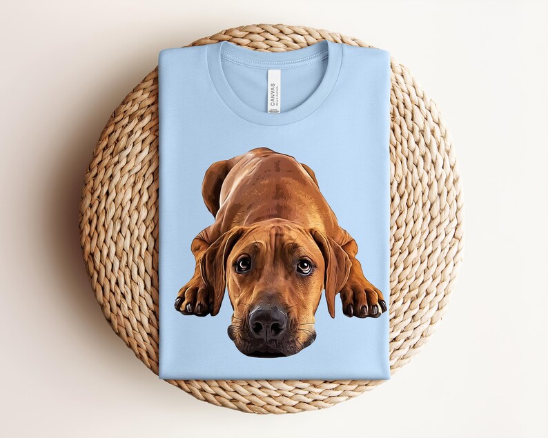 May include: A light blue t-shirt featuring a centered, detailed illustration of a brown dog in a lying position. The dog has large, expressive eyes, floppy ears, and a black nose. The shirt has a crew neckline and a white brand label at the neck.