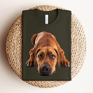 May include: A dark olive green t-shirt featuring a realistic graphic illustration of a brown Rhodesian Ridgeback dog lying down in a prone position. The dog has large, expressive eyes and a black nose, centered on the front of the shirt.