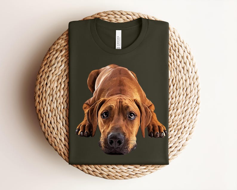 May include: A dark olive green t-shirt featuring a realistic graphic illustration of a brown Rhodesian Ridgeback dog lying down in a prone position. The dog has large, expressive eyes and a black nose, centered on the front of the shirt.