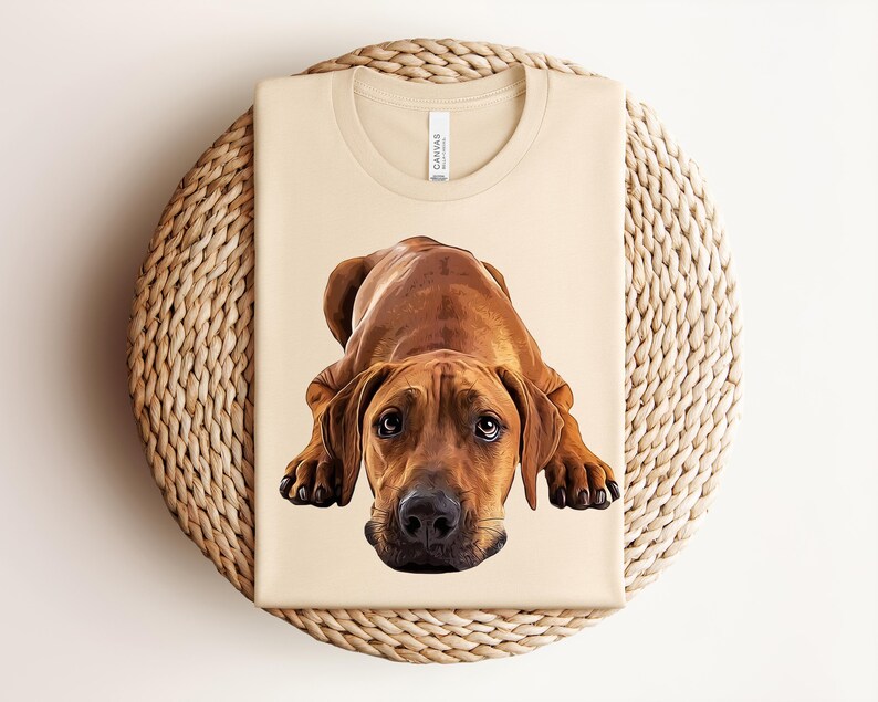 May include: A cream-colored t-shirt featuring a detailed, realistic illustration of a brown Rhodesian Ridgeback dog lying down in a prone position. The dog has dark brown eyes, a black nose, and short fur. The shirt has a crew neckline.