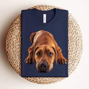 May include: A navy blue t-shirt featuring a graphic illustration of a brown dog lying down with its head facing forward. The dog has large, expressive eyes and a black nose. The shirt has a crew neckline and a white brand label at the collar.