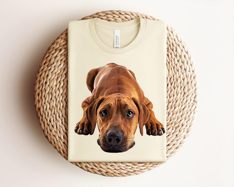 May include: A cream-colored crew neck t-shirt featuring a detailed, realistic illustration of a brown Rhodesian Ridgeback dog lying down in a playful, forward-facing position with its head resting on the ground and dark eyes looking upward.