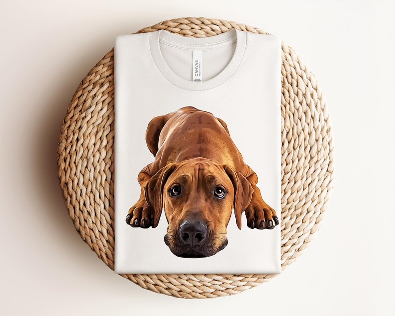 May include: A white short-sleeved t-shirt featuring a detailed, realistic illustration of a brown Rhodesian Ridgeback dog lying down in a prone position. The dog has dark eyes, a black nose, and floppy ears, centered on the front of the shirt.