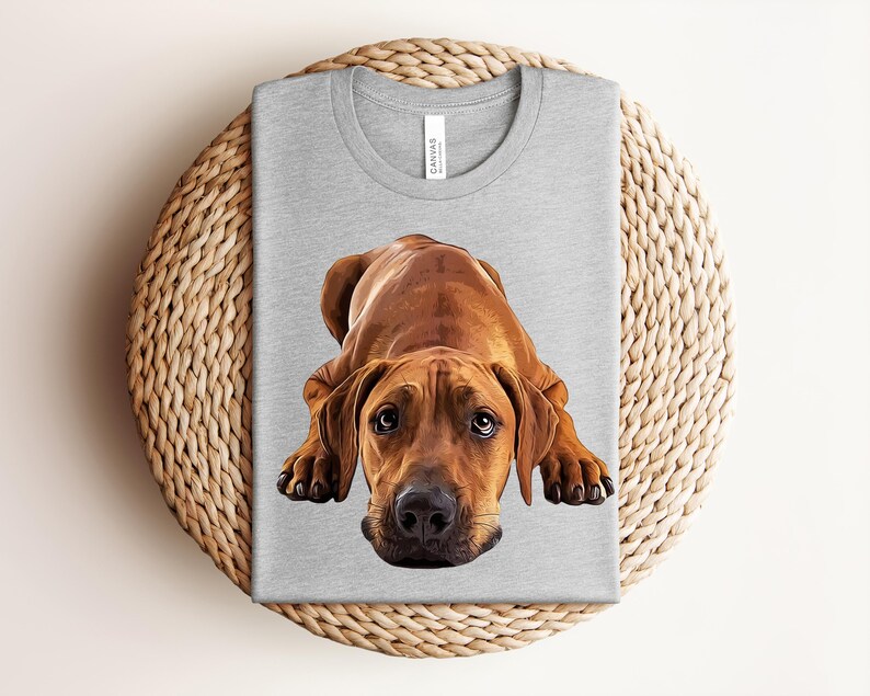 May include: A light heather gray crew neck t-shirt featuring a detailed, realistic illustration of a brown dog with floppy ears and dark eyes lying down in a resting position. The dog is centered on the front of the shirt, facing forward.