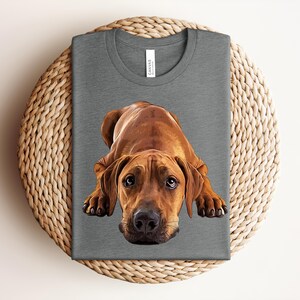 May include: A heather gray crew neck t-shirt featuring a detailed, realistic illustration of a brown Rhodesian Ridgeback dog lying down with its head facing forward. The dog has dark eyes, a black nose, and floppy ears, centered on the shirt.