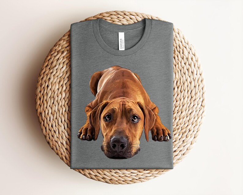 May include: A heather gray crew neck t-shirt featuring a detailed, realistic illustration of a brown Rhodesian Ridgeback dog lying down with its head facing forward. The dog has dark eyes, a black nose, and floppy ears, centered on the shirt.