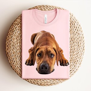 May include: A light pink t-shirt featuring a detailed, realistic illustration of a brown Rhodesian Ridgeback dog lying down in a resting position. The dog has dark eyes, a black nose, and floppy ears, centered on the front of the garment.