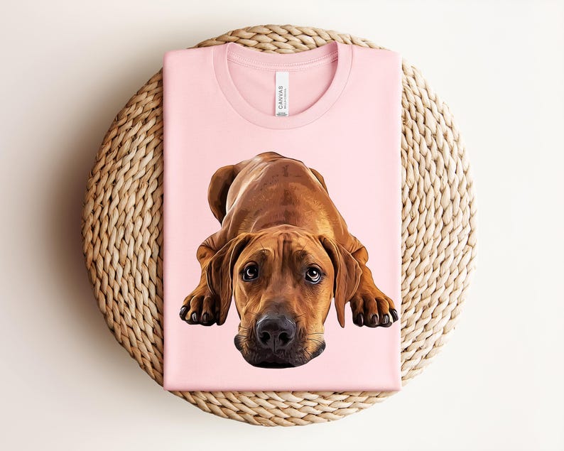 May include: A light pink t-shirt featuring a detailed, realistic illustration of a brown Rhodesian Ridgeback dog lying down in a resting position. The dog has dark eyes, a black nose, and floppy ears, centered on the front of the garment.