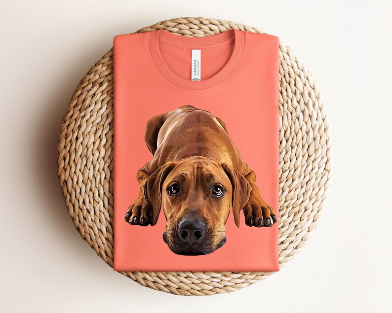 May include: A coral-colored crew neck t-shirt featuring a detailed graphic illustration of a brown dog lying down in a prone position. The dog has large, expressive eyes and a black nose. The shirt has a white label inside the collar area.